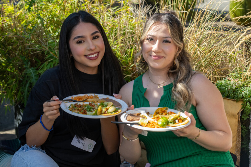 Two women sitting together, smiling, holding a plate of food at a Morrison appreciation event.